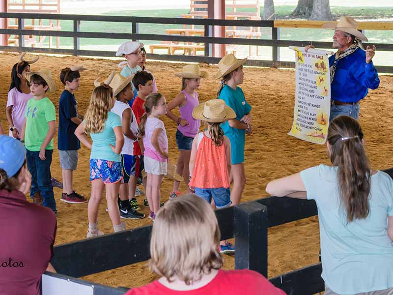 Children in horse arena with Man in Cowboy hat holding sign